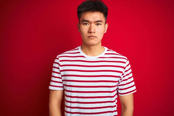 孤立した赤い背景の上に立つストライプのTシャツを着た若いアジアの中国人男性は 真剣な表情でリラックスしました カメラを見てシンプルで自然な — ストック写真