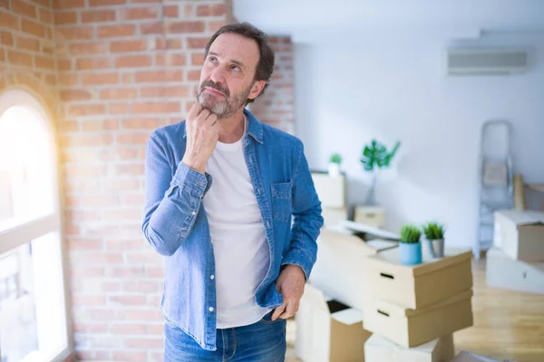 Middle age senior man moving to a new house packing cardboard boxes with hand on chin thinking about question, pensive expression. Smiling with thoughtful face. Doubt concept.