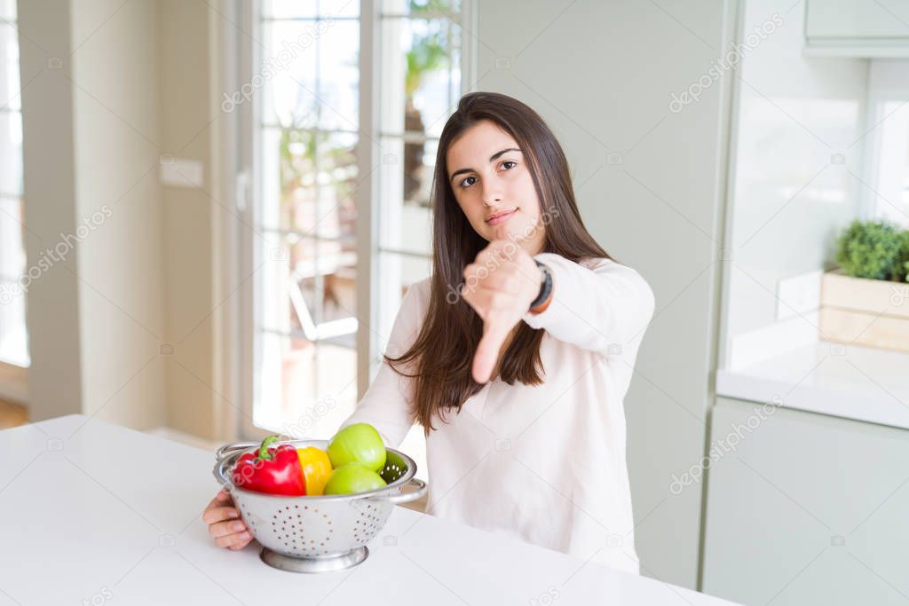 Beautiful young woman using colander to wash and clean vegatables with angry face, negative sign showing dislike with thumbs down, rejection concept