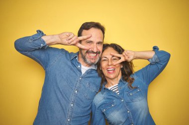 Güzel orta yaş çift birlikte izole sarı arka plan üzerinde ayakta yüz üzerinde parmaklar ile Barış sembolü yapıyor, neşeli gösteren zaferle gülümseyen