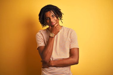 Dreadlocks ile Afro adam izole sarı arka plan üzerinde duran çizgili t-shirt ayakta silah ve el çene üzerinde yükseltilmiş ile gülümseyen kamera emin bakıyor. Pozitif düşünme.