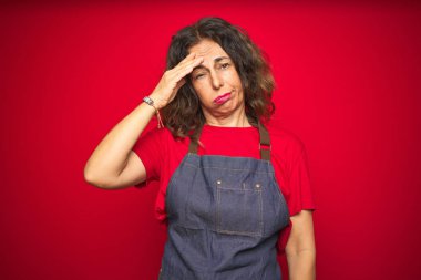 Kırmızı izole arka plan üzerinde apron üniforma giyen orta yaş kıdemli kadın endişeli ve alnında el, sinir ve kriz için endişeli bir sorun hakkında vurguladı
