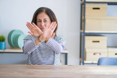 Orta yaş kıdemli kadın evde masada oturan ret ifadesi geçiş silah ve avuç içi negatif işareti, öfkeli yüz yapıyor