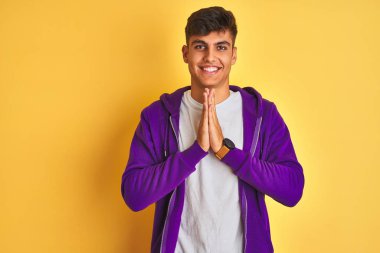 Mor sweatshirt giyen genç hintli adam izole sarı arka plan üzerinde duran elleri birlikte emin gülümseyerek af isteyen dua.