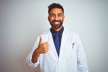 Genç Hint Doktor adam ayakta üzerinde izole beyaz arka plan yapıyor mutlu thumbs yukarı jest ile el. Başarı göstererek kameraya bakarak ifadeyi onaylama.