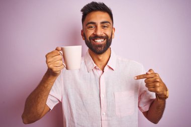 Genç Hint adam giyen gömlek içme fincan arasında kahve ayakta üzerinde izole pembe arka plan ile sürpriz yüz işaret parmak için kendisi