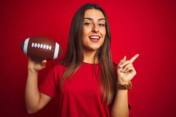 年轻美丽的女运动员拿着足球在孤立的红色背景很高兴指着手和手指的一边 — 图库照片