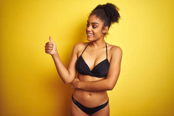 African american woman on vacation wearing bikini standing over isolated yellow background Looking proud, smiling doing thumbs up gesture to the side