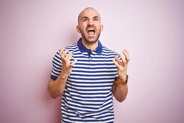 Young bald man with beard wearing casual striped blue t-shirt over pink isolated background crazy and mad shouting and yelling with aggressive expression and arms raised. Frustration concept.