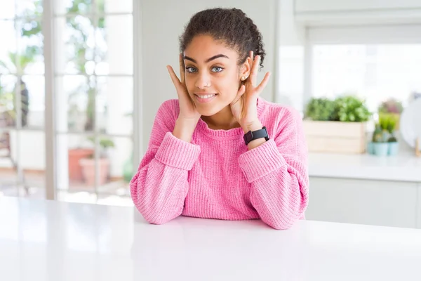 Beautiful african american woman with afro hair wearing casual pink sweater Trying to hear both hands on ear gesture, curious for gossip. Hearing problem, deaf