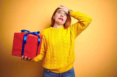 Sarı izole arka plan üzerinde tutarak Doğum günü hediyesi tutan genç güzel Redhead kadın utanç ve sürpriz yüz, öfkeli ve sinirli ile şok, baş el ile vurguladı. Korku ve hata için üzgün.