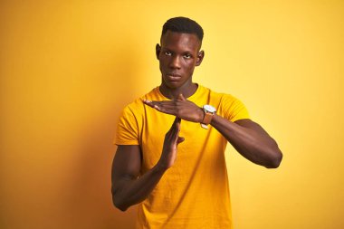 Young african american man wearing casual t-shirt standing over isolated yellow background Doing time out gesture with hands, frustrated and serious face clipart