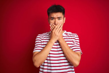 İzole kırmızı arka plan üzerinde ayakta çizgili t-shirt duran Genç Asyalı Çinli adam yanlışlıkla eller ile ağız kapsayan şok. Gizli konsept.