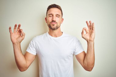 Casual beyaz t-shirt izole arka plan üzerine dinlenmek giydiği ve gözleri ile gülümseyen yakışıklı delikanlı parmak ile meditasyon jest yapıyor kapalı. Yoga kavramı.