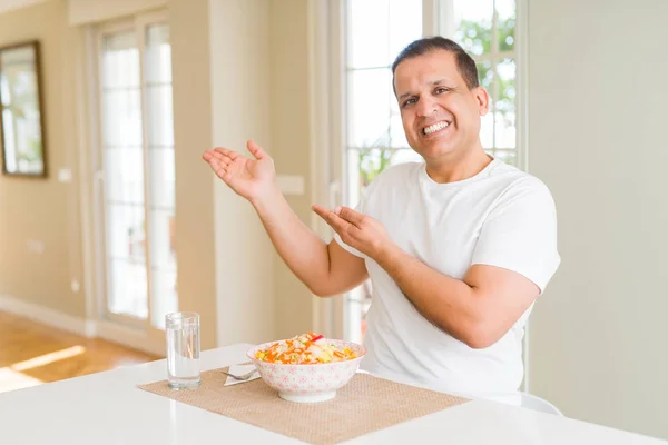 Middle age man eating rice at home amazed and smiling to the camera while presenting with hand and pointing with finger.