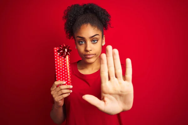 Young african american woman holding valentine gift over isolated red background with open hand doing stop sign with serious and confident expression, defense gesture