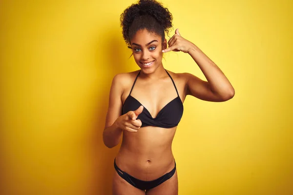 African american woman on vacation wearing bikini standing over isolated yellow background smiling doing talking on the telephone gesture and pointing to you. Call me.