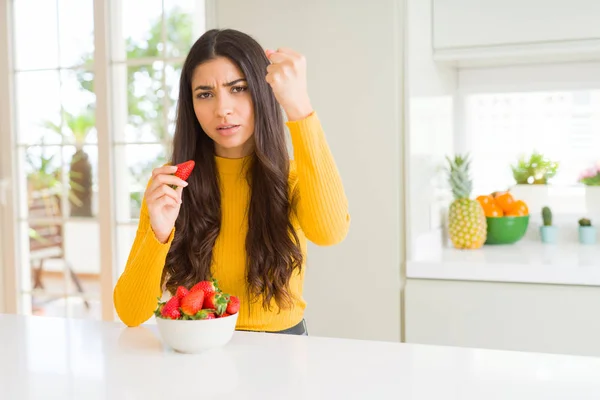 Young woman eating fresh red strawberry as healthy snack annoyed and frustrated shouting with anger, crazy and yelling with raised hand, anger concept