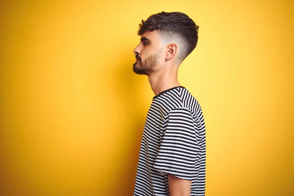 Young man with tattoo wearing striped t-shirt standing over isolated yellow background looking to side, relax profile pose with natural face with confident smile.