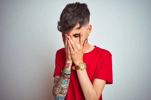 Young man with tattoo wearing red t-shirt standing over isolated white background with sad expression covering face with hands while crying. Depression concept.