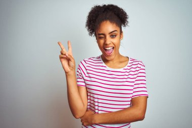 Afrika Amerikan kadın rahat pembe çizgili t-shirt üzerinde izole beyaz arka plan üzerinde mutlu yüz kırmızı fotoğraf makinesi zafer işareti yapıyor ile gülümsüyor giyiyor. İki numara.