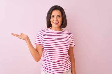 İzole pembe arka plan üzerinde duran çizgili t-shirt giyen genç güzel kadın neşeli sunan gülümseyerek ve kameraya bakarak avuç içi ile işaret.
