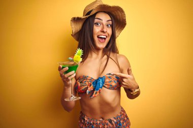 Kadın tatil üzerinde bikini ve şapka içme kokteyl üzerinde izole sarı arka plan çok mutlu el ve parmak ile işaret