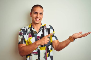 Beyaz izole arka plan üzerinde yaz gömlek giyen genç yakışıklı adam hayran ve kamera gülümseyen el ile sunmak ve parmak ile işaret.