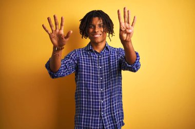İzole sarı arka plan üzerinde ayakta sıradan gömlek giyen dreadlocks ile Afro adam gösteren ve parmak numarası dokuz ile işaret kadar emin ve mutlu gülümseyen.