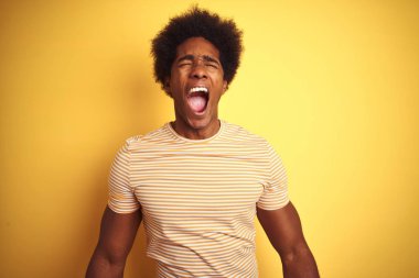 Afro saç ile Amerikan adam izole sarı arka plan üzerinde duran çizgili t-shirt ayakta kızgın ve deli çığlık sinirli ve öfkeli, öfke ile bağırıyor. Öfke ve agresif kavram.