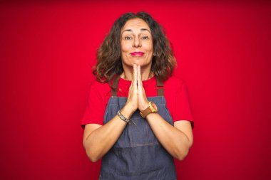 Orta yaş kıdemli kadın kırmızı izole arka plan üzerinde apron üniforma giyen el ile birlikte affetmek için güven gülümseyen soran dua.