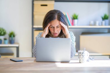 Orta yaş kıdemli kadın baş ağrısı çaresizlikten muzdarip bilgisayar laptop kullanarak evde çalışan masada oturan ve ağrı ve migren nedeniyle vurguladı. Eller baş.