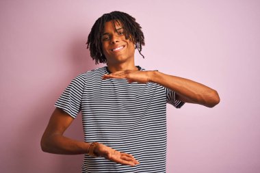 Siyah büyük ve büyük boyutlu işareti, ölçmek sembolü gösteren eller ile izole pembe arka plan üzerinde ayakta lacivert çizgili t-shirt giymiş dreadlocks ile Afro adam. Kameraya bakarak gülümsüyor. Ölçme konsepti.