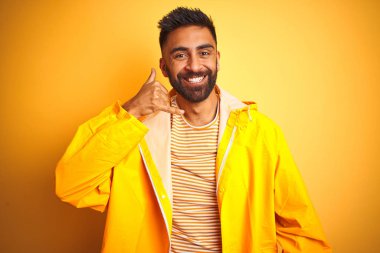 Genç Hint adam cep telefonda konuşmak gibi el ve parmaklar ile telefon jest yaparken gülümseyen izole sarı arka plan üzerinde ayakta yağmurluk giyiyor. İletişim kavramları.