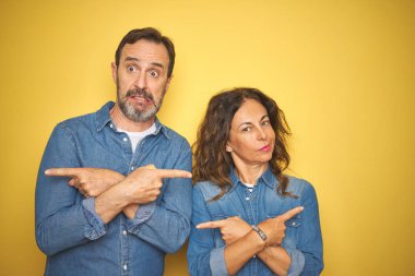 Güzel orta yaş çift birlikte izole sarı arka plan üzerinde ayakta parmaklarla her iki tarafa Işaret, farklı yönde katılmıyorum