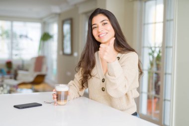 Genç kadın evde bir fincan kahve içme mutlu büyük gülümseme ile ok işareti, başparmak yukarı parmaklar, mükemmel işareti yapıyor