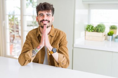 Genç adam giyen Casual ceket beyaz masa üzerinde oturan eller ile birlikte affetmek için yalvarıyor güven gülümseyen.