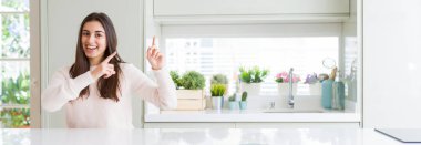 Güzel genç kadın evde beyaz masaya oturan geniş açı resim gülümseyen ve iki elleri ve parmakları ile tarafına işaret kamera bakıyor.