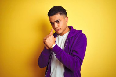 İzole sarı arka planda ayakta mor sweatshirt giyen genç Brezilyalı adam el jest ile sembolik silah Holding, öldürme çekim silah oynarken, öfkeli yüz