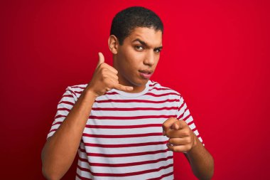 İzole kırmızı arka plan üzerinde çizgili t-shirt giyen genç yakışıklı Arap adam telefon jest işeyerek ve sizi işaret gülümseyerek. Beni ara.