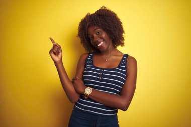 Yüzünde büyük bir gülümseme ile izole sarı arka plan üzerinde çizgili t-shirt giyen genç Afrikalı Afro kadın, kamera bakarak yan el ve parmak ile işaret.