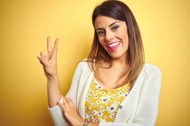 Sarı izole arka plan üzerinde ayakta ceket giyen genç güzel kadın mutlu yüz zafer işareti yapıyor kamera göz kırpıyor ile gülümsüyor. İki numara.