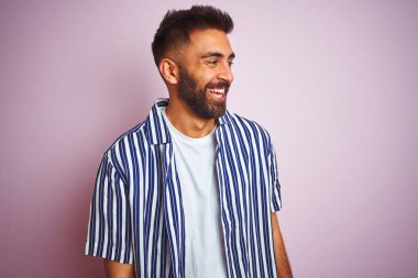 Genç yakışıklı Hint adam giyen yaz çizgili gömlek üzerinde izole pembe arka plan üzerinde yüz gülümseme ile yan bakıyor, doğal ifade. Güven gülüyor.