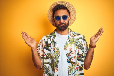 Hint adam tatil giyen çiçek gömlek şapka gözlük üzerinde izole sarı arka plan clueless ve karıştı ifade ile silah ve eller kaldırdı. Şüphe kavramı.