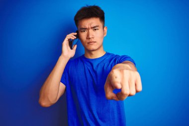 Genç Asyalı Çinli adam kamera ve size, el işareti, önde pozitif ve emin jest parmak ile izole mavi arka plan üzerinde ayakta akıllı telefon üzerinde konuşuyor