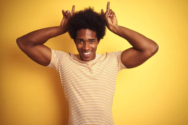 American man with afro hair wearing striped t-shirt standing over isolated yellow background Posing funny and crazy with fingers on head as bunny ears, smiling cheerful