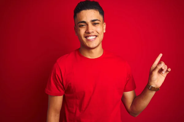 孤立した赤い背景の上に立つTシャツを着た若いブラジル人男性は カメラを見て手指で指差し 顔に大きな笑みを浮かべて — ストック写真