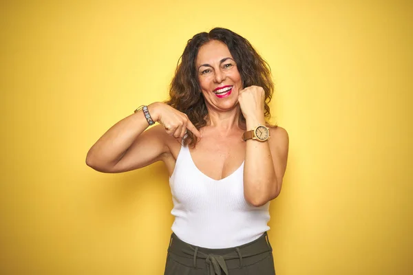 Middle age senior woman showing wrist watch over yellow isolated background with surprise face pointing finger to himself