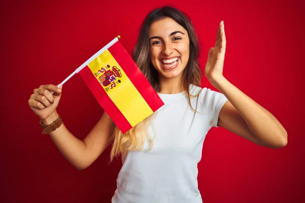 年轻的美丽女人拿着西班牙国旗在红色孤立的背景非常高兴和兴奋 胜利者的表情庆祝胜利尖叫与大大的微笑和举手 — 图库照片