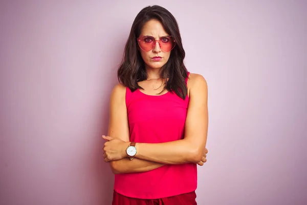 Beautiful woman wearing funny pink heart shaped sunglasses over pink isolated background skeptic and nervous, disapproving expression on face with crossed arms. Negative person.